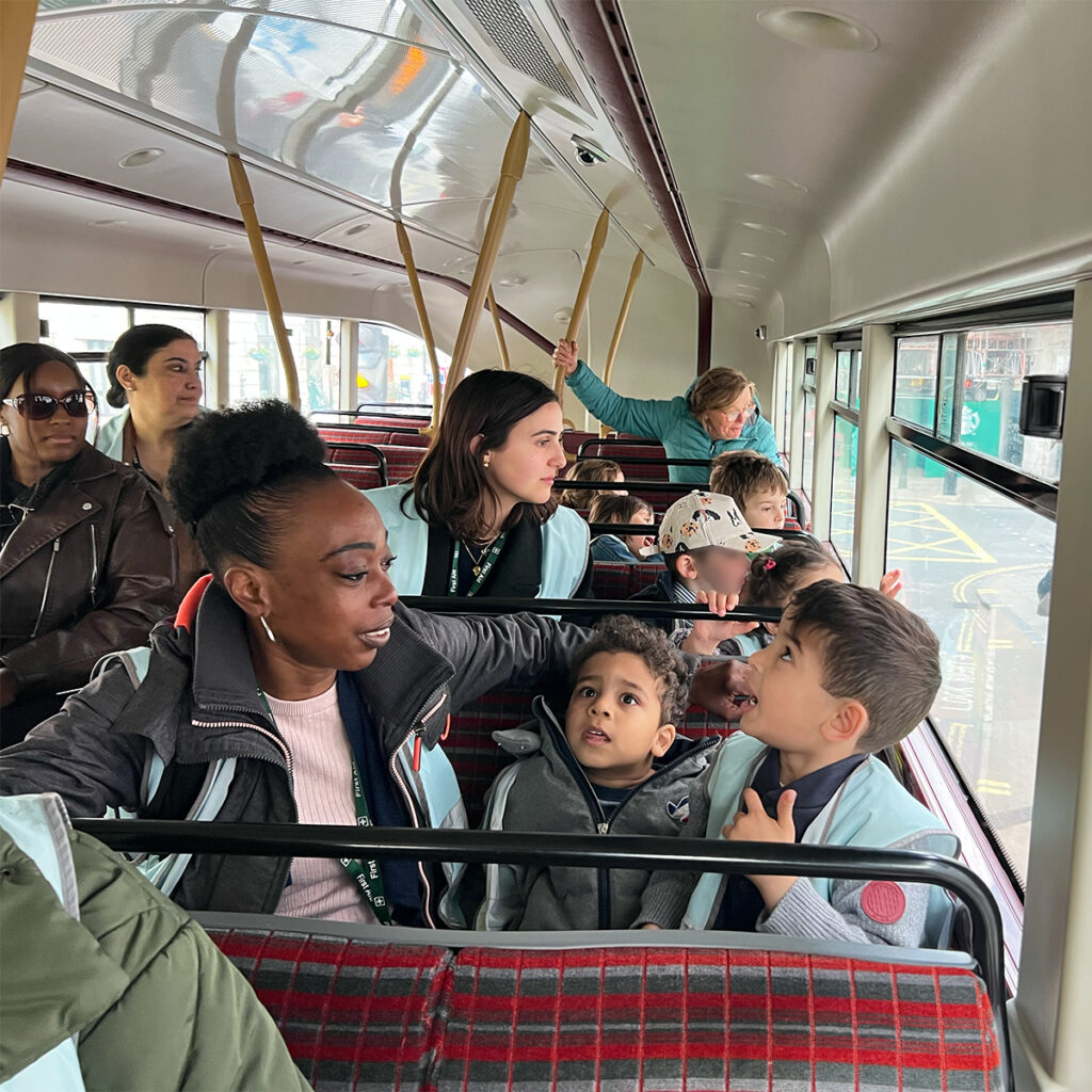 EIFA A group of young children and adults wearing coats and name tags are seated on a bus. Two children are conversing animatedly, while a few adults look on. The bus has red plaid seats, and some passengers hold onto overhead straps.