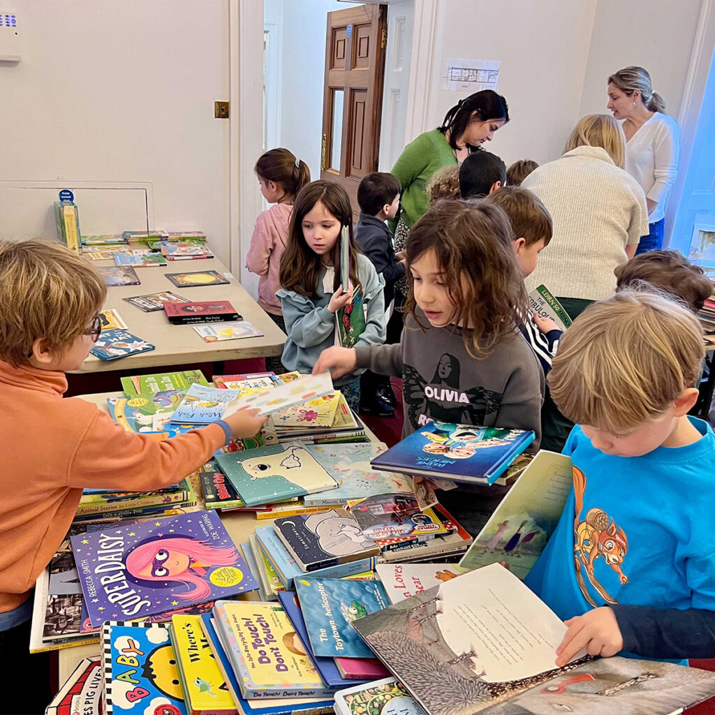 EIFA Les enfants se rassemblent autour de tables remplies de livres jeunesse colorés. Certains feuilletent, d'autres lisent ou discutent. Des adultes les assistent. L'ambiance est animée et stimulante, évoquant un salon du livre ou un événement de lecture.
