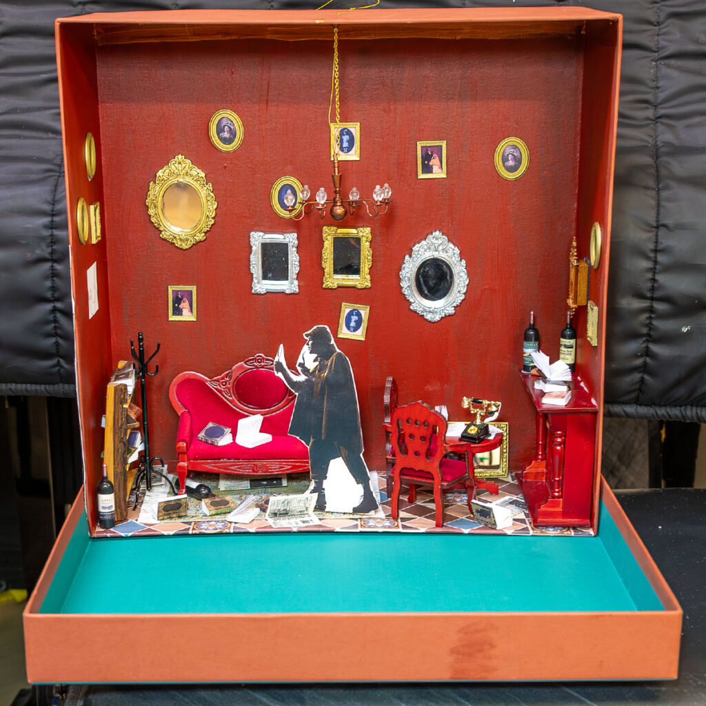 EIFA Diorama d'une pièce vintage aux murs rouges, avec un canapé rouge et des papiers éparpillés. Une silhouette représentant une personne debout en train de lire. La pièce est ornée de cadres dorés, d'un lustre, d'une petite table et d'une bouteille de vin. Un sol bleu contraste avec la décoration chaleureuse.